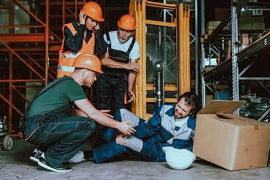 İş kazasında çalışan hakları ve destek süreçleri İş kazası geçiren bir çalışana yardım eden ekip, iş güvenliği ekipmanlarıyla dolu bir depo ortamında.