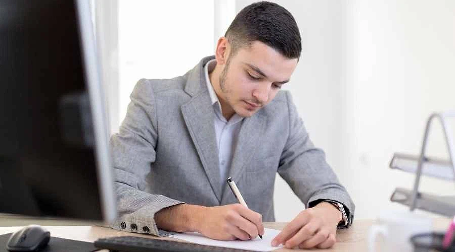 2026 memur maaşları güncel bilgileri Ofis ortamında, gri takım elbise giymiş bir erkek, masa başında yazı yazıyor.