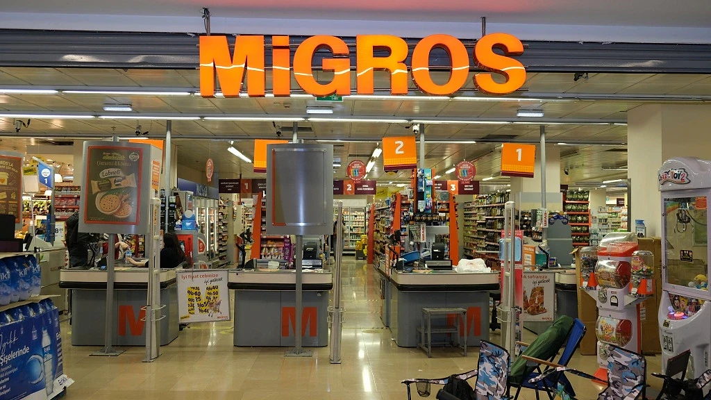 Migros maaşları hakkında detaylı bilgi Migros marketinin iç görünümü, kasalar ve ürün reyonlarıyla dolu.