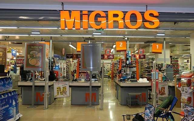 Migros maaşları hakkında detaylı bilgi Migros marketinin iç görünümü, kasalar ve ürün reyonlarıyla dolu.