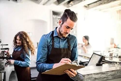 Saatlik çalışma avantajları ve şartları hakkında bilgi Kafe ortamında çalışan bir adam, not alırken. Arka planda başka çalışanlar var.