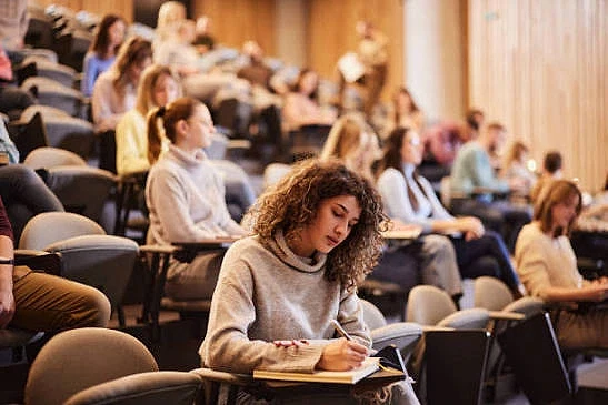 Konferans salonunda katılımcılar ve not alan genç kadın Bir konferans salonunda not alan genç kadın, arka planda katılımcılar dikkatle dinliyor.