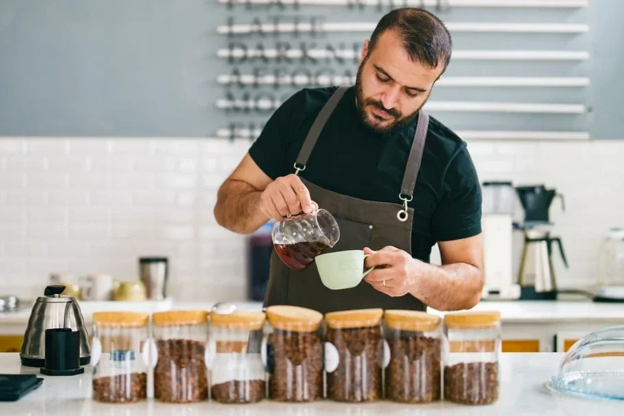 Güncel Part Time Barista İş İlanları ve Rehberi Kahve hazırlayan barista, kahve çekirdekleriyle dolu kavanozların önünde çalışıyor.